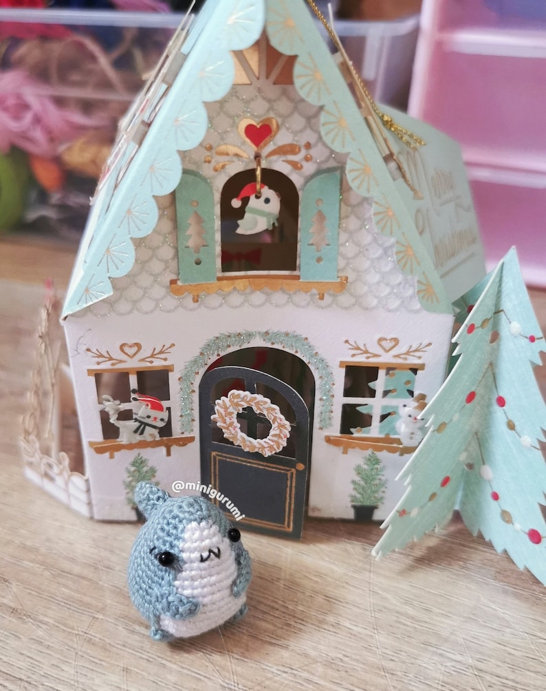 Pu&ograve; includere: Una piccola statuina di pinguino in crochet blu e bianco si trova davanti a una casa di Natale in carta con una ghirlanda sulla porta e un albero di Natale a destra. La casa &egrave; decorata con glitter e ha un piccolo uccello in una finestra.