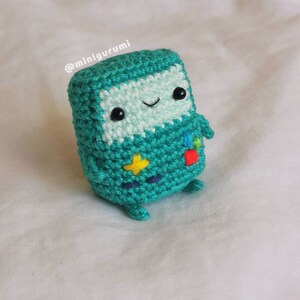 May include: A teal blue crocheted plush toy with a smiling face, black eyes, and a white mouth. The toy has a yellow star, red square, and blue circle on its body. The toy is on a white surface. @minigurumi