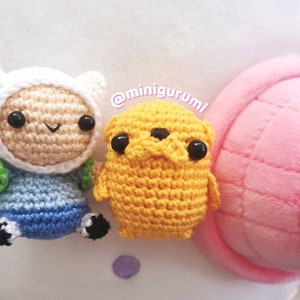 May include: Crocheted Finn the Human and Jake the Dog from Adventure Time. Finn is wearing a white hat and blue and green clothing. Jake is yellow and has black eyes. The characters are sitting on a pink and white surface.