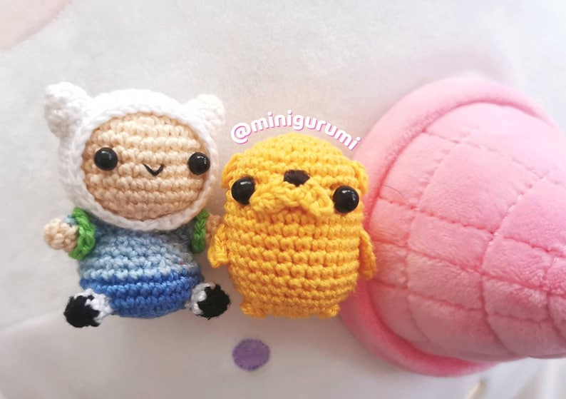 May include: Crocheted Finn the Human and Jake the Dog from Adventure Time. Finn is wearing a white hat and blue and green clothing. Jake is yellow and has black eyes. The characters are sitting on a pink and white surface.