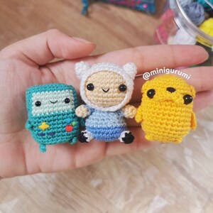 May include: Three crocheted stuffed toys: a green Finn the Human, a blue and white Jake the Dog, and a yellow and brown BMO. The toys are sitting on a hand. The text '@minigurumi' is in the background.