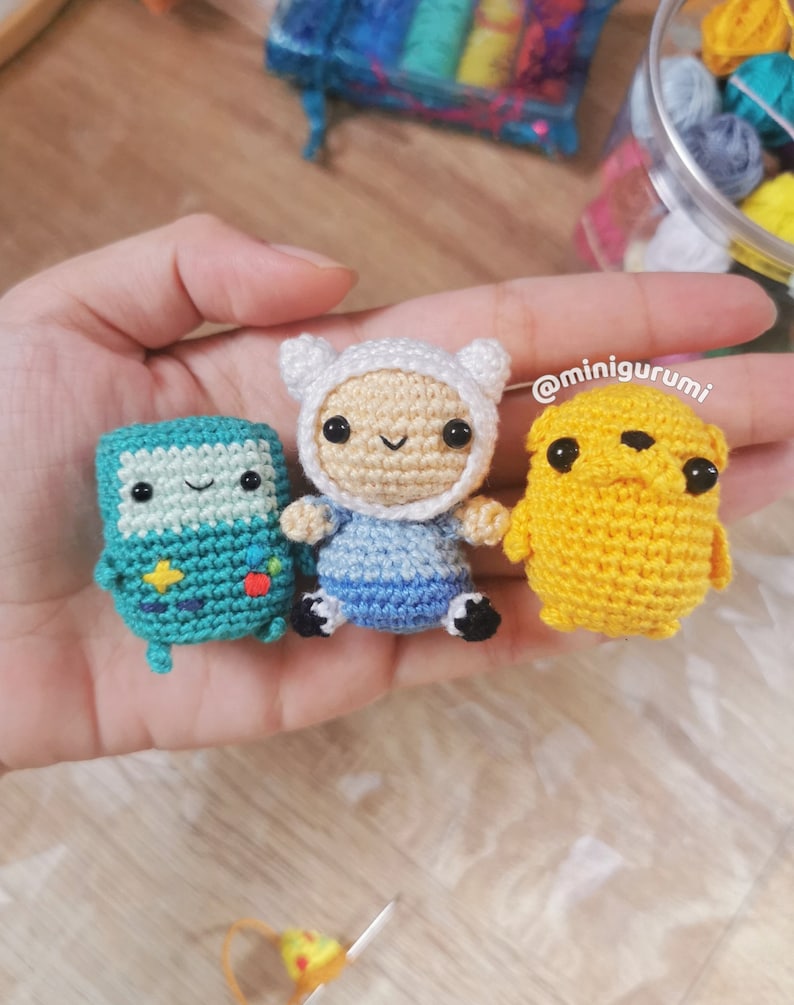 May include: Three crocheted stuffed toys: a green Finn the Human, a blue and white Jake the Dog, and a yellow and brown BMO. The toys are sitting on a hand. The text '@minigurumi' is in the background.