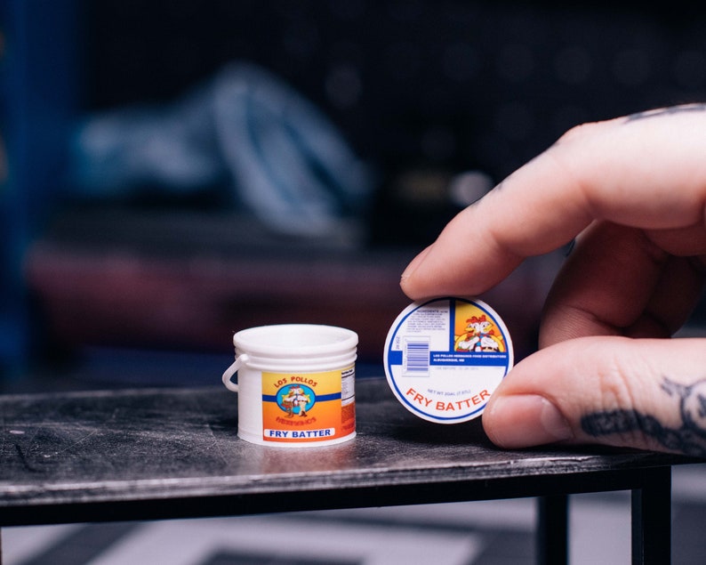 May include: Miniature white bucket labeled "Los Pollos Fry Batter" with a yellow, orange, and blue design. A round lid with the same design is held nearby. The items are on a dark surface.