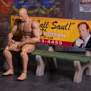May include: A tan action figure sits on a green bench with gray supports. Behind the figure is a sign that says "Call Saul!" with a picture of a man pointing. The sign is yellow and red. The background is a gray wall with graffiti.