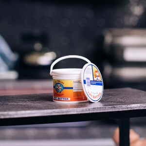 May include: A white plastic bucket with a handle and a lid, labeled "Los Pollos Hermanos Fry Batter." The label features a cartoon image of a chef. The bucket sits on a dark surface.