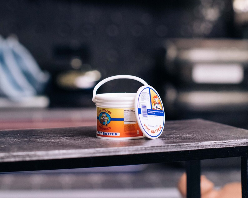 May include: A white plastic bucket with a handle and a lid, labeled "Los Pollos Hermanos Fry Batter." The label features a cartoon image of a chef. The bucket sits on a dark surface.