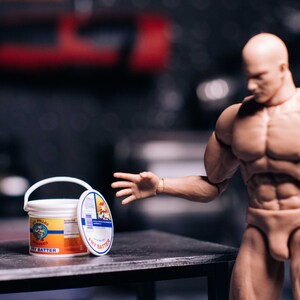 May include: A white bucket of "Fry Batter" with a red and orange label, and a lid with a blue and white label. A muscular action figure is reaching towards the bucket on a dark surface.