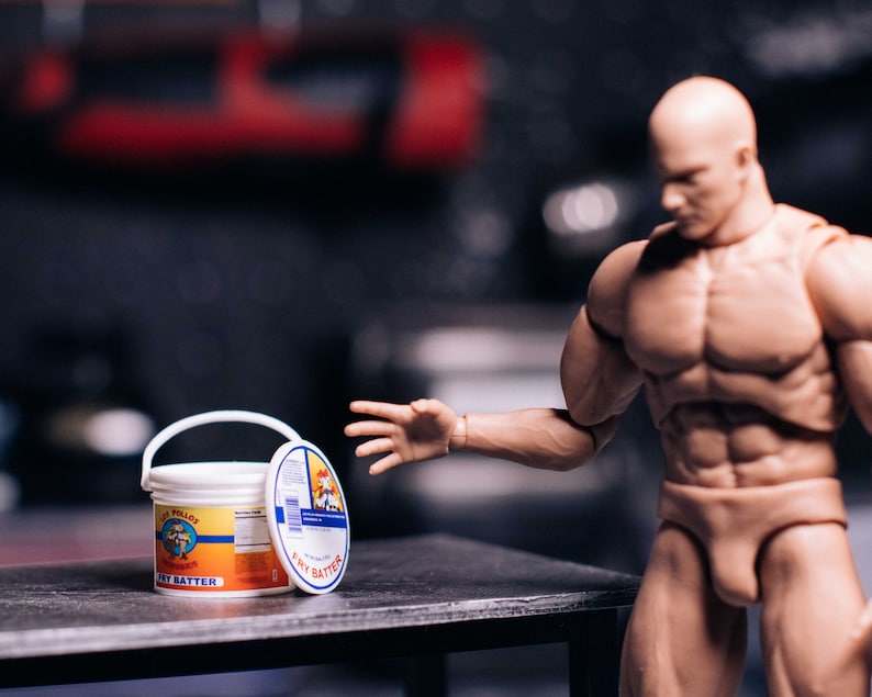 May include: A white bucket of "Fry Batter" with a red and orange label, and a lid with a blue and white label. A muscular action figure is reaching towards the bucket on a dark surface.