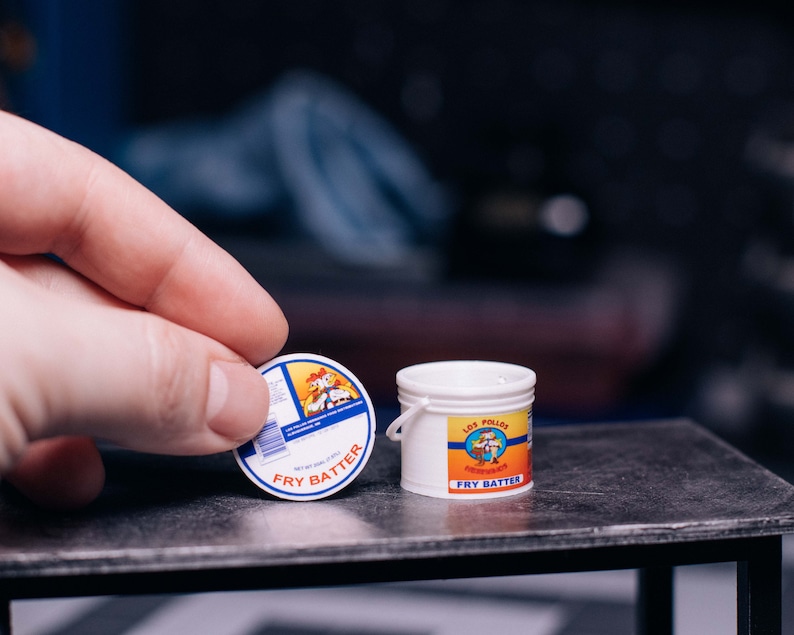 May include: Miniature white bucket and a round label with the text "FRY BATTER". The label features a cartoon chicken and the words "LOS POLLOS HERMANOS". The bucket has a small handle. The items are on a dark surface.