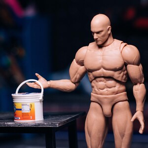 May include: A muscular action figure with a bald head and tan skin, holding a white bucket with a blue and orange label. The figure is posed on a dark surface, with a blurred background.