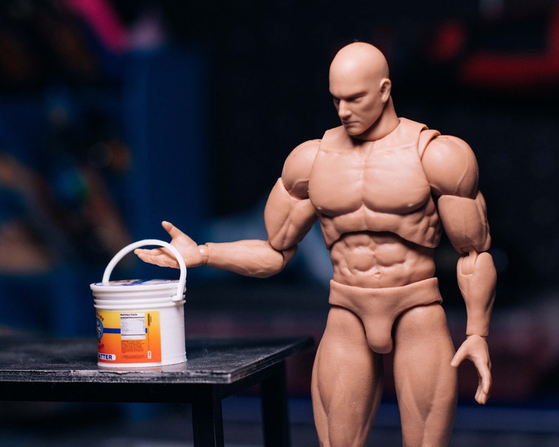 May include: A muscular action figure with a bald head and tan skin, holding a white bucket with a blue and orange label. The figure is posed on a dark surface, with a blurred background.