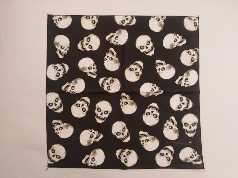 May include: A square bandana featuring a repeating pattern of off-white human skulls on a solid black background. The skulls are oriented in various directions across the fabric, displaying detailed eye sockets, nasal cavities, and teeth.