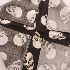 May include: A fabric cloth featuring a repeating pattern of off-white skulls with dark eye sockets and red pupils set against a textured charcoal gray background. The material is folded into a square shape with visible black stitched edges.