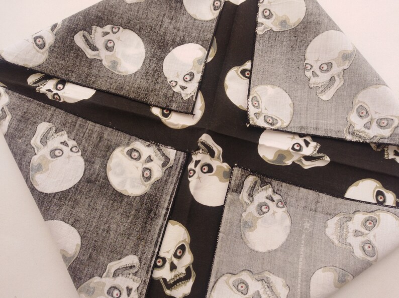 May include: A fabric cloth featuring a repeating pattern of off-white skulls with dark eye sockets and red pupils set against a textured charcoal gray background. The material is folded into a square shape with visible black stitched edges.