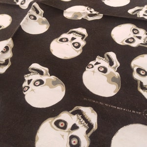 May include: A black fabric featuring a repeating pattern of off-white skulls with dark eye sockets and red pupils. The fabric includes printed text that reads 50% COTTON 50% POLY HAV-A-HANK MADE IN U.S.A. RN 15187 along with a small star logo.