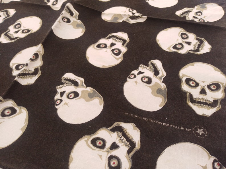 May include: A black fabric featuring a repeating pattern of off-white skulls with dark eye sockets and red pupils. The fabric includes printed text that reads 50% COTTON 50% POLY HAV-A-HANK MADE IN U.S.A. RN 15187 along with a small star logo.