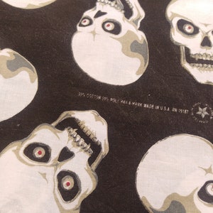 May include: A black fabric featuring a repeating pattern of off-white skulls with dark eye sockets and red pupils. The fabric includes printed text: 50% COTTON 50% POLY HAV-A-HANK MADE IN U.S.A. RN 15187, along with a star logo containing the text CRAFTED WITH PRIDE.