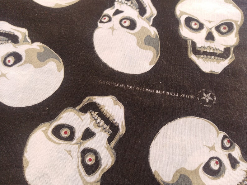 May include: A black fabric featuring a repeating pattern of off-white skulls with dark eye sockets and red pupils. The fabric includes printed text: 50% COTTON 50% POLY HAV-A-HANK MADE IN U.S.A. RN 15187, along with a star logo containing the text CRAFTED WITH PRIDE.