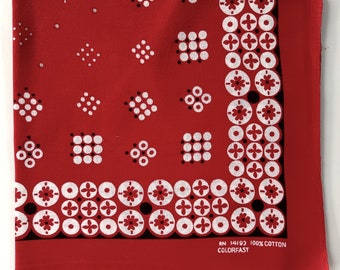 Pode incluir: Bandana vermelha com padrões geométricos brancos e pretos. A borda apresenta um design floral repetitivo. O texto "RN 14193 100% COTTON COLORFAST" está impresso no canto inferior direito.