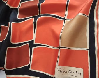 May include: A square scarf featuring a geometric grid pattern of orange, dark red, and tan squares outlined in white against a black background. The fabric has a smooth, satin-like finish. A white rectangular label with the text Pierre Cardin is visible.