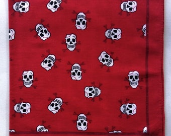 Puede incluir: Un pañuelo cuadrado de color rojo con un patrón repetitivo de calaveras blancas con detalles en negro y huesos cruzados. El borde inferior incluye el texto impreso: 100% COTTON MADE IN CHINA COLORFAST RN 13881. La tela tiene un tejido visible y un borde rojo oscuro.
