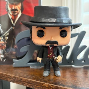 以下が含まれることがあります： 黒い帽子、黒いスーツ、赤いベストを着たキャラクターのFunko Popビニールフィギュア。フィギュアは口ひげを生やしており、小さな銃を持っています。キャラクターの額縁入り画像が背景にあります。