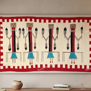Puede incluir: Un tapiz rectangular con fondo crema y borde rojo. El diseño presenta figuras estilizadas con detalles en negro, rojo y gris, y faldas azules. La obra de arte se exhibe sobre un mueble de madera.