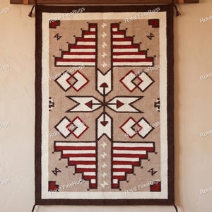 Op de afbeelding: Een rechthoekig wandkleed met een geometrisch patroon in tinten bruin, donkerbruin, wit en dieprood. Het ontwerp bevat trapvormige motieven, pijlfiguren en kleine kruissymbolen, omlijst door een dikke donkerbruine rand met kwastjes aan de hoeken.