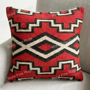 Op de afbeelding: Een vierkant decoratief kussen met een geometrisch patroon in rood, zwart en wit. Het kussen ligt op een lichtgrijze fauteuil. Het patroon bevat zigzags en ruitvormen.