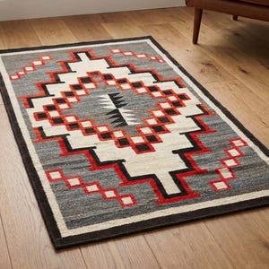 Pode incluir: Tapete retangular cinza com um padrão geométrico em vermelho, branco e preto. O tapete tem uma borda escura e está colocado sobre um piso de madeira. Uma pequena planta em vaso e uma poltrona de couro estão no fundo.