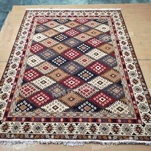 May include: Rectangular area rug with a geometric diamond pattern in shades of red, brown, beige, and blue. The rug has a detailed border with a repeating design and fringe along the edges. The rug is displayed on a light brown surface.