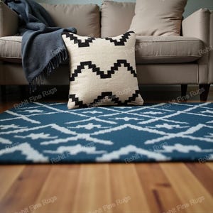 Op de afbeelding: Een beige bank met een blauwe deken en een decoratief kussen met een zwart geometrisch patroon. Een blauw tapijt met een wit geometrisch ontwerp ligt op de houten vloer. De afbeelding toont texturen en patronen.