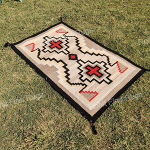 May include: Rectangular rug with a geometric pattern in beige, black, red, and gray. The rug has a black border with tassels at each corner. The central design features a cross-like shape in red.