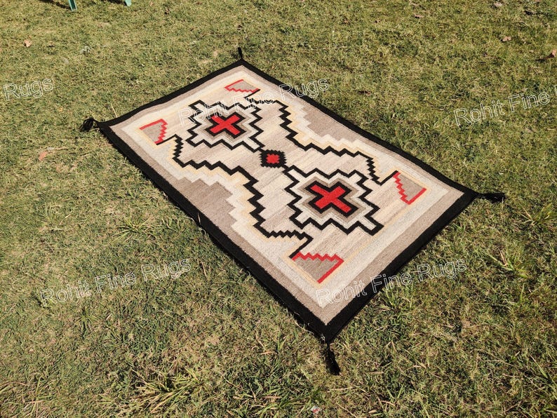 May include: Rectangular rug with a geometric pattern in beige, black, red, and gray. The rug has a black border with tassels at each corner. The central design features a cross-like shape in red.