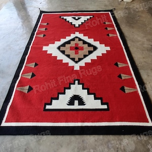 Pode incluir: Um tapete retangular vermelho com um padrão geométrico em branco, preto e castanho. O tapete tem uma borda preta e borlas decorativas nos cantos. O design central apresenta uma forma de diamante com uma cruz.