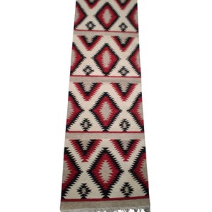 Puede incluir: Una alfombra rectangular con un patrón geométrico. La alfombra presenta un fondo color crema con patrones repetidos de diamantes y zig-zag en rojo, negro y gris. Los bordes tienen flecos.