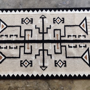 Può includere: Un tappeto beige con un motivo geometrico bianco e nero. Il tappeto ha un bordo nero con una frangia bianca e nera.