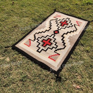 May include: Rectangular woven rug with a geometric design. The rug features a central pattern with red crosses and black zigzag lines on a beige background. The border is black, and the rug is displayed on green grass.