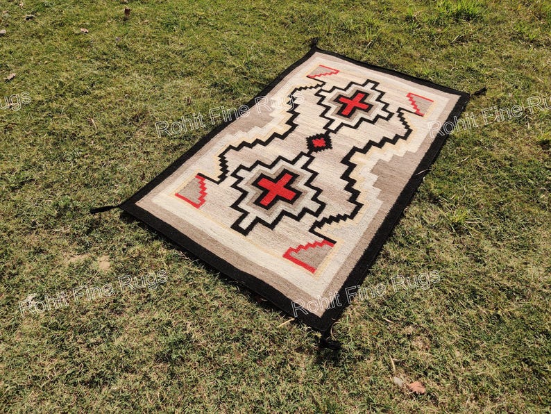 May include: Rectangular woven rug with a geometric design. The rug features a central pattern with red crosses and black zigzag lines on a beige background. The border is black, and the rug is displayed on green grass.
