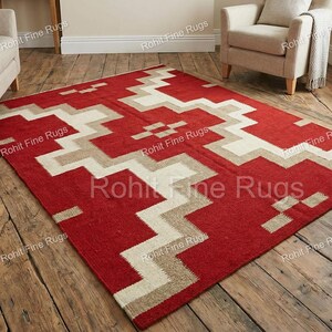 May include: A rectangular red rug with a geometric pattern in beige and white. The rug is placed on a wooden floor, and the design features zig-zag and rectangular shapes. The rug is a home decor item.