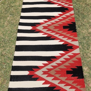Puede incluir: Una alfombra larga y estrecha con un fondo a rayas blanco y negro y un patrón geométrico rojo, blanco y marrón en un lado. La alfombra está hecha de lana y tiene un diseño tradicional nativo americano.