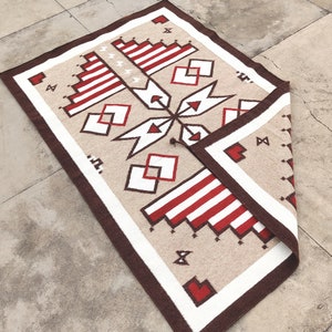 Può includere: Un tappeto beige con un motivo geometrico rosso e bianco. Il tappeto ha un bordo marrone ed è piegato a metà.