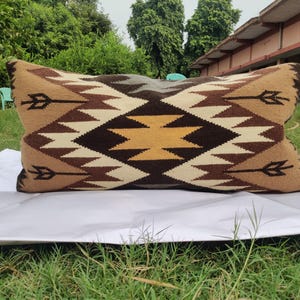 May include: Rectangular decorative pillow with a geometric pattern in shades of brown, tan, and cream. The pillow features a central diamond design with arrow-like motifs. The pillow is placed on a white surface against a backdrop of green grass and trees.