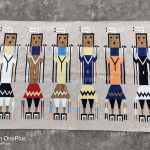 May include: Rectangular beige rug with six stylized figures in various colors. Each figure has a rectangular head, a patterned torso, and a skirt-like bottom. The figures are arranged in a row, with the rug's edges finished with tassels.