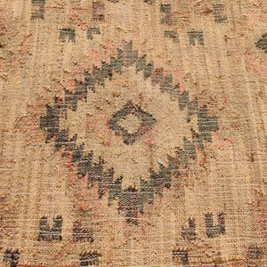 Puede incluir: Primer plano de una alfombra beige tejida con un patr&oacute;n geom&eacute;trico. El dise&ntilde;o presenta una forma de diamante central y motivos repetidos en negro y rosa. La alfombra tiene una superficie texturizada.