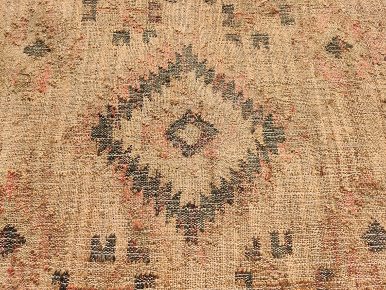 Puede incluir: Primer plano de una alfombra beige tejida con un patr&oacute;n geom&eacute;trico. El dise&ntilde;o presenta una forma de diamante central y motivos repetidos en negro y rosa. La alfombra tiene una superficie texturizada.