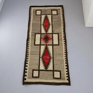 May include: A rectangular, woven rug with a geometric design. The rug features a central pattern of red diamonds and brown squares on a gray and cream background. The border is dark brown with a zigzag pattern.