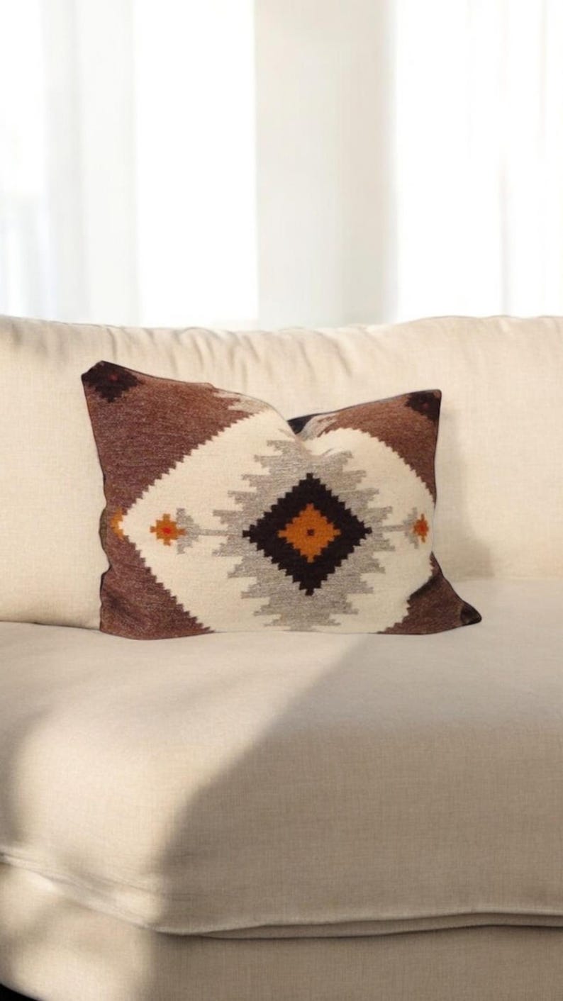 Pu&ograve; includere: Cuscino decorativo con motivo geometrico. Il cuscino presenta un motivo a diamante centrale nei toni del marrone, grigio e arancione su sfondo bianco. &Egrave; posto su un divano color crema.