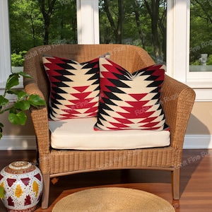 Op de afbeelding: Twee decoratieve sierkussens met een geometrisch patroon in rood, zwart en crème. De kussens liggen op een rieten stoel met een crèmekleurig kussen. De stoel staat voor een raam met uitzicht op bomen.