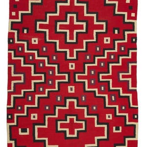 Op de afbeelding: Een rechthoekig, rood geweven tapijt met een geometrisch patroon. Het ontwerp toont witte, zwarte en paarse trapvormige lijnen en kruisvormen tegen een rode achtergrond. Het tapijt heeft een gefranjerde rand.